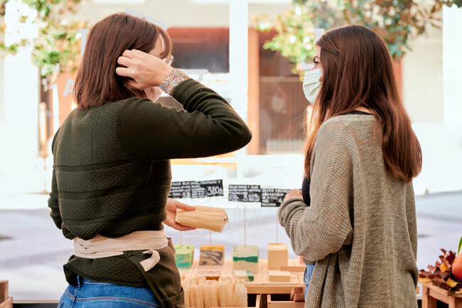 Back view of female buyer in medical mask talking to saleswoman while standing near shelf with organic soap — Stock Photo