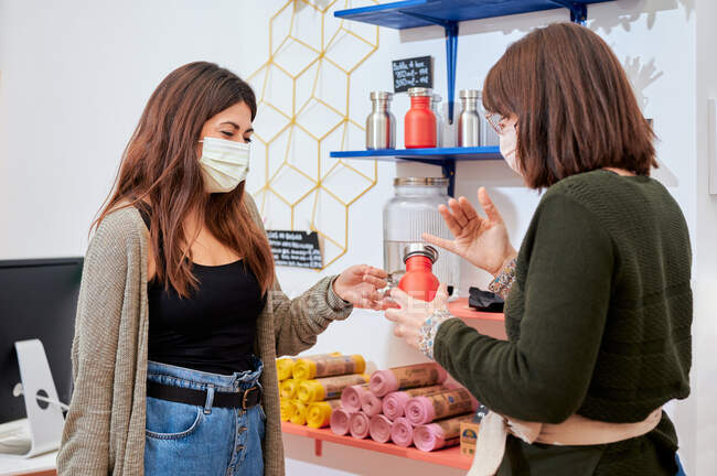 Продавец женского пола в защитной маске консультирует клиента по выбору экологически чистой металлической бутылки в магазине — стоковое фото