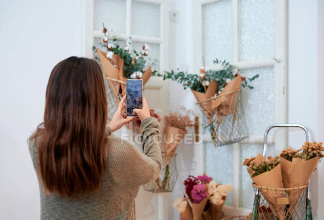 Visão traseira de uma fêmea irreconhecível tirando fotos de flores embrulhadas em papel vendendo em loja a granel com produtos ecológicos — Fotografia de Stock