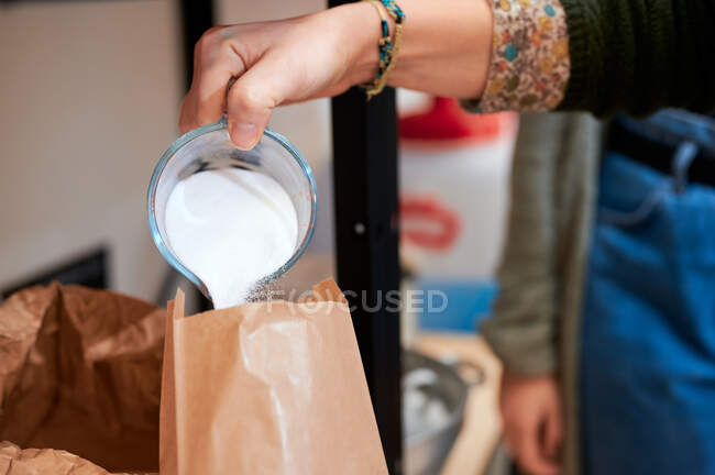 Vendedora anónima de cultivos vertiendo polvo de lavado en peso del vaso de precipitados en el paquete de papel mientras hace el pedido para el cliente en la tienda a granel - foto de stock