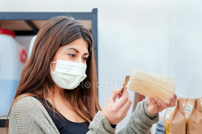 Vista lateral do cliente feminino em máscara médica ler informações sobre etiqueta de preço de papel durante as compras em loja a granel — Fotografia de Stock