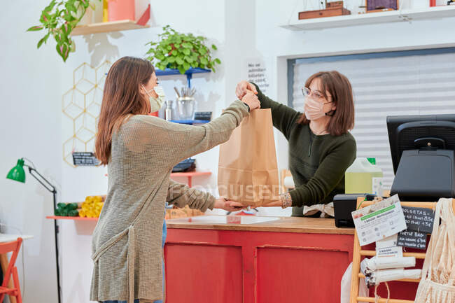 Frau mit Schutzmaske gibt Kundin Papiereinkaufstüte, während sie am Schalter im Großmarkt arbeitet — Stockfoto