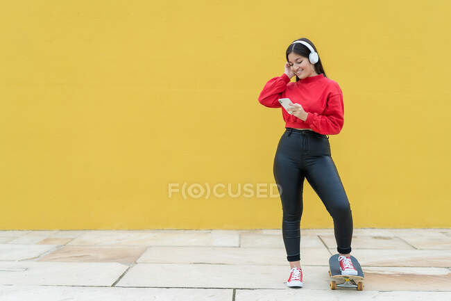 Молодая веселая этническая спортсменка слушает музыку из наушников, наблюдая за сотовым телефоном и стоя на улице со скейтбордом на желтом фоне — стоковое фото