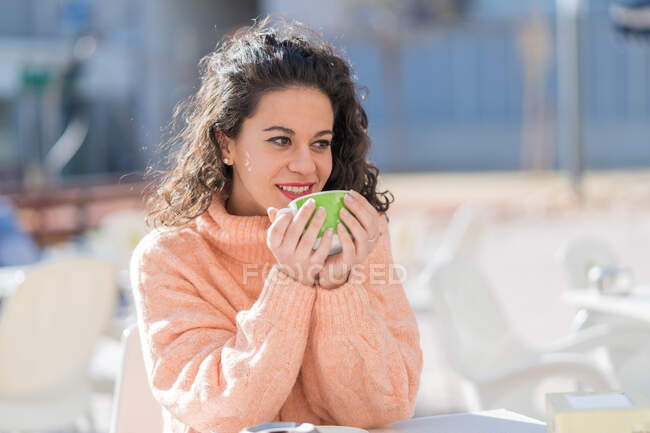 Affascinante giovane bruna indossa maglione rosa sciolto godendo di caffè appena fatto e guardando via contento mentre si siede a tavola in un caffè all'aperto soleggiato — Foto stock