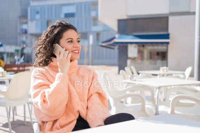 Delighted smiling brunette in cozy pink sweater talking on mobile phone on sunny outdoor cafe — Stock Photo