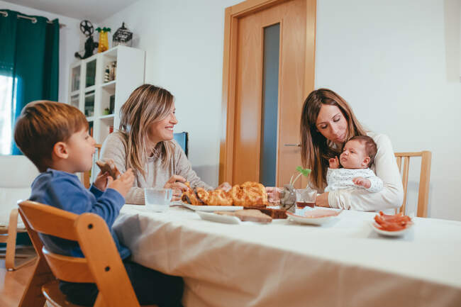 Mulher homossexual alegre com pequeno bebê sentado à mesa com namorada e filho enquanto tendo refeição — Fotografia de Stock