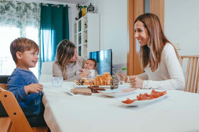Mulher homossexual alegre com pequeno bebê sentado à mesa com namorada e filho enquanto tendo refeição — Fotografia de Stock