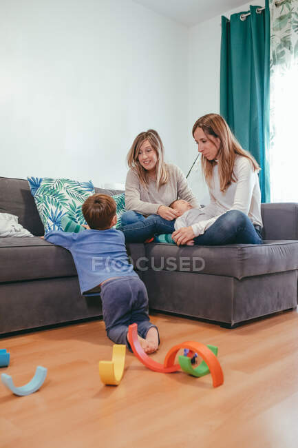 Comprimento completo alegre jovem casal lésbico em casual desgaste descansando no sofá aconchegante e olhando para o filho bonito jogando no chão — Fotografia de Stock