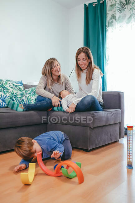 Полностью веселая молодая лесбийская пара в повседневной одежде отдыхает на уютном диване и смотрит на милого сына, играющего на полу — стоковое фото