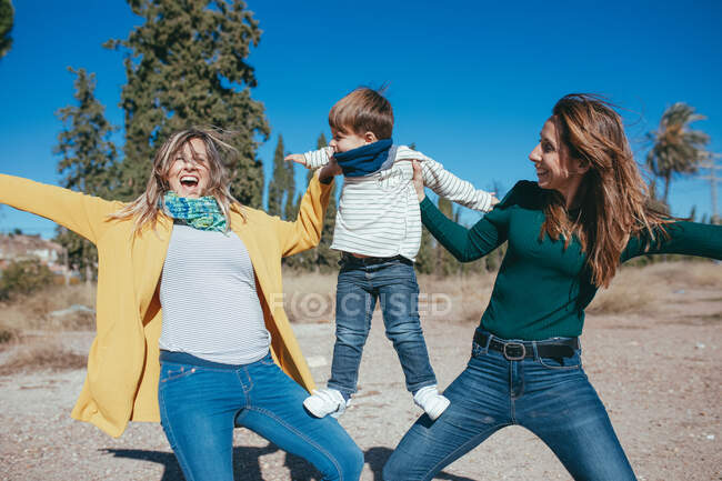 Joyful young lesbian couple wearing casual clothes lifting adorable little son on hands while spending time together in sunny nature — Stock Photo