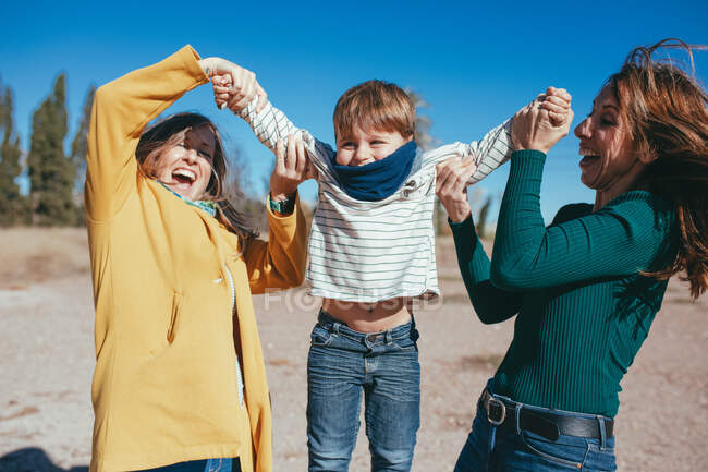 Alegre joven lesbiana pareja usando casual ropa lifting adorable pequeño hijo en manos mientras pasando tiempo juntos en sunny naturaleza - foto de stock