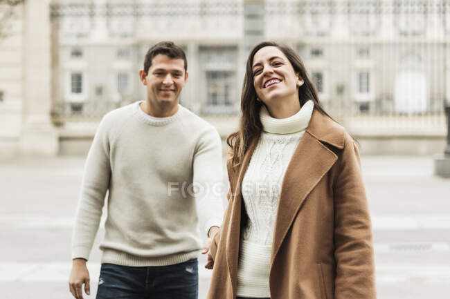 Cheerful young couple wearing warm clothes holding hands while walking together on asphalt sidewalk in modern city — Stock Photo