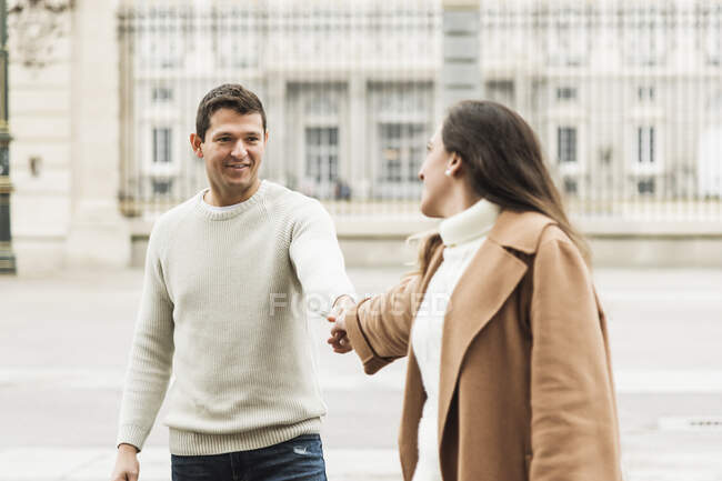 Jovem casal alegre vestindo roupas quentes de mãos dadas enquanto caminham juntos na calçada de asfalto na cidade moderna — Fotografia de Stock