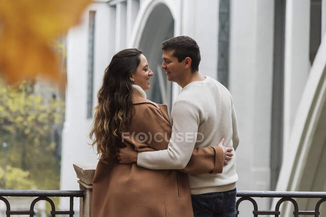 Vista posterior alegre pareja joven en ropa de abrigo abrazándose tiernamente y mirándose con afecto mientras están de pie juntos en la calle - foto de stock
