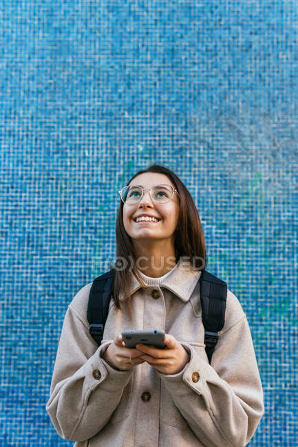 Урожай приваблива усміхнена студентка-жінка в теплому одязі з рюкзаком, використовуючи сучасний мобільний телефон на тротуарі на вулиці — стокове фото