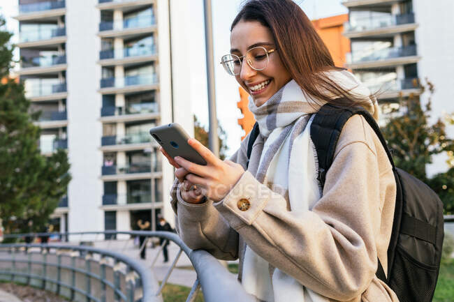 Cultiver attrayant sourire étudiante en tenue chaude avec sac à dos en utilisant un téléphone portable moderne sur le trottoir dans la rue — Photo de stock