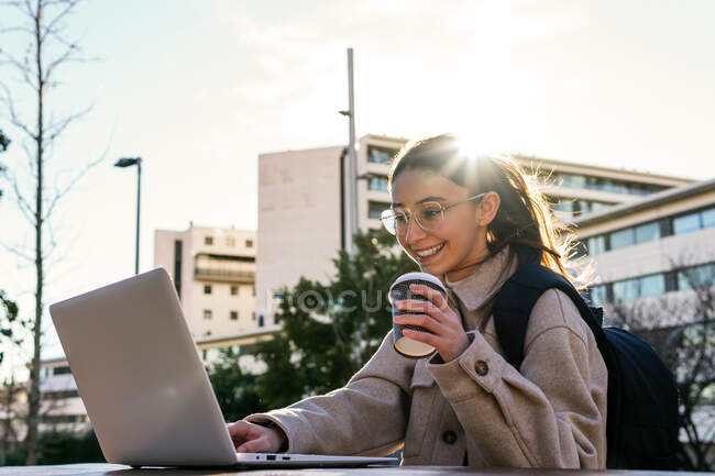Feliz estudiante sonriente con abrigo cálido y anteojos con mochila trabajando en asignación a domicilio en netbook mientras está sentada en la mesa con café para llevar en el soleado parque - foto de stock