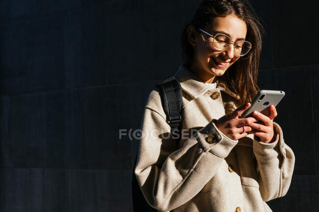 Обрезание улыбается студентка в теплом пальто и очки с рюкзаком просматривает мобильный телефон, стоя на солнечной улице тротуар — стоковое фото