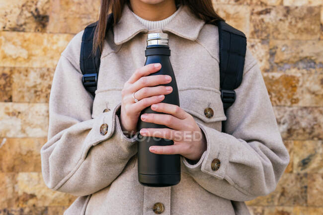 Crop young female wearing warm coat with reusable bottle while standing on street on autumn day — Stock Photo