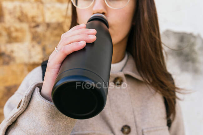 Colheita jovem do sexo feminino vestindo casaco quente água potável de garrafa reutilizável preto, enquanto em pé na rua no dia de outono — Fotografia de Stock