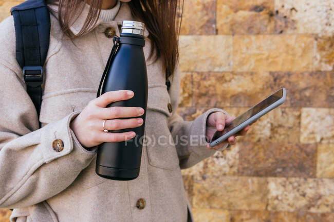 Crop anonymous female in warm coat with backpack holding reusable water bottle and browsing modern mobile phone against building wall on street — Stock Photo