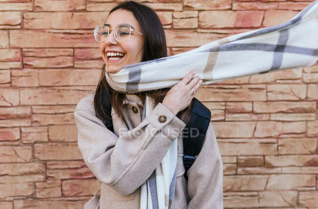 Emocionado joven mujer feliz en abrigo cálido y gafas con mochila poniéndose una bufanda voladora acogedora mientras está de pie en la pasarela de la calle - foto de stock