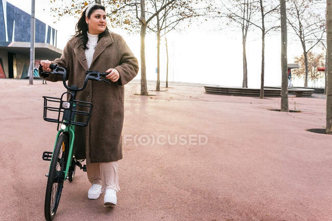 Joven hembra gordita contemplativa en abrigo paseando con bicicleta contra árboles otoñales mientras mira hacia otro lado en la ciudad - foto de stock