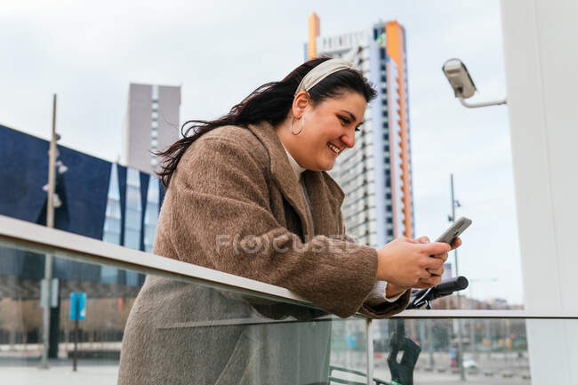 Вид сбоку на молодую весёлую пухлую женщину в пальто, болтающую по мобильному телефону, опираясь на забор в городе — стоковое фото
