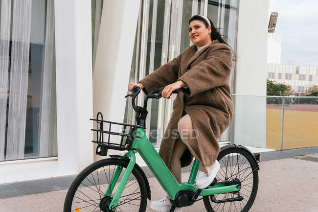 Молода споглядальна пухка жінка в пальто, що йде з велосипедом проти осінніх дерев, дивлячись далеко в місто — стокове фото