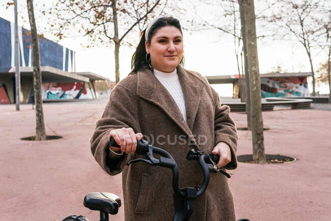 Jeune femme potelée contemplative en manteau se promenant à vélo contre les arbres d'automne tout en regardant loin en ville — Photo de stock