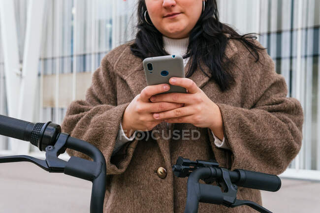 Crop vista di giovane femmina grassoccio allegro in cappotto chattare sul cellulare mentre si appoggia in città — Foto stock