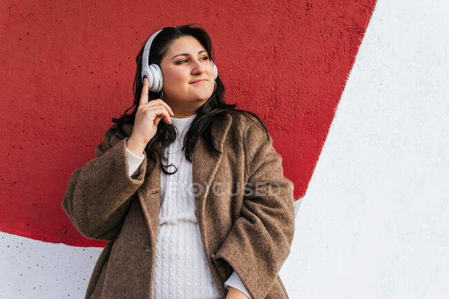 Молодая мечтательная пухленькая женщина в пальто слушает песню в беспроводных наушниках, глядя на улицу — стоковое фото