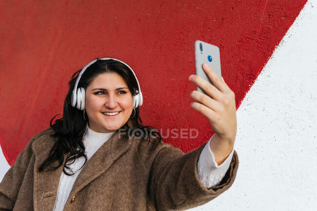 Молодая веселая женщина с избыточным весом делает автопортрет на сотовом телефоне, слушая музыку в беспроводных наушниках возле яркой стены — стоковое фото