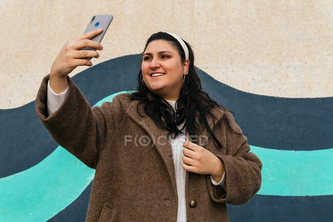 Giovane allegra femmina sovrappeso prendendo autoritratto sul telefono cellulare vicino alla parete luminosa — Foto stock