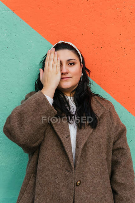 Young overweight female in coat and headband covering eye while looking at camera in daylight — Stock Photo