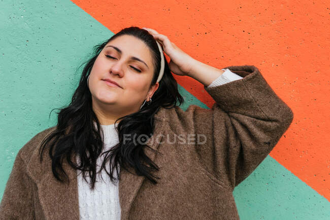Jovem mulher com sobrepeso em casaco e cabeça tocando cabeça com olhos fechados à luz do dia — Fotografia de Stock