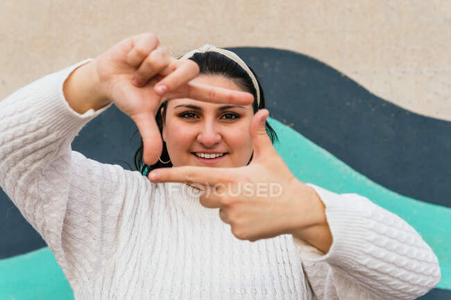 Young content plump female in white knitwear demonstrating frame gesture while looking at camera near wall — Stock Photo