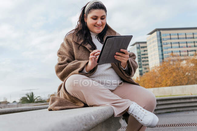 Молода усміхнена пухнаста жінка в повсякденному одязі переглядає Інтернет на планшеті, відпочиваючи на лавці проти міської будівлі восени — стокове фото
