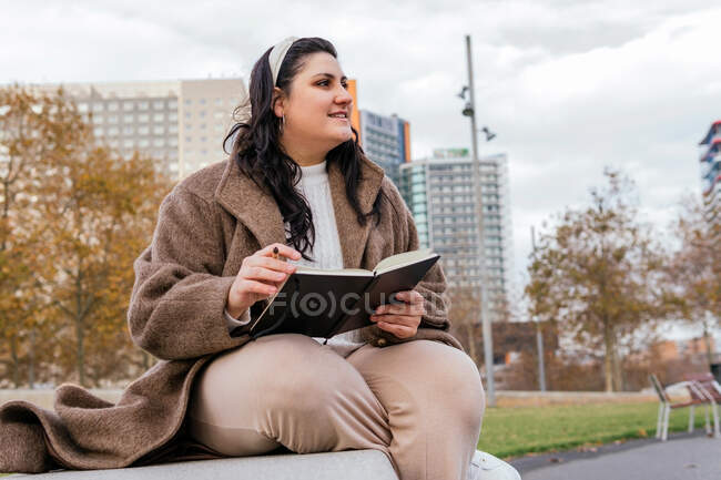 Joven mujer regordeta sonriente en ropa casual escribiendo notas en un cuaderno mientras descansa en el banco contra el edificio urbano en otoño - foto de stock