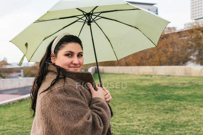 Vue latérale de la jeune femme dodue sincère en manteau avec parapluie regardant la caméra le jour de pluie — Photo de stock