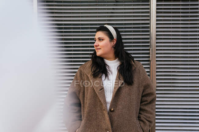 Side view of young contemplative plus size female with hand in pocket looking away under cloudy sky in city — Stock Photo