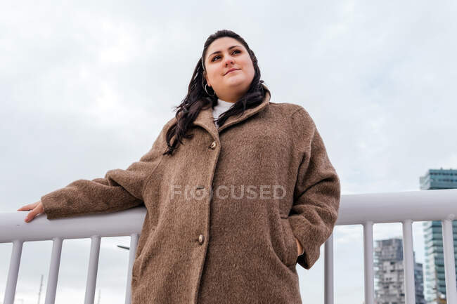 From below of young contemplative plus size female with hand in pocket looking away under cloudy sky in city — Stock Photo