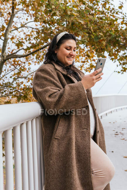 Young glad chubby female in coat surfing internet on mobile phone while leaning against a fence in town — Stock Photo