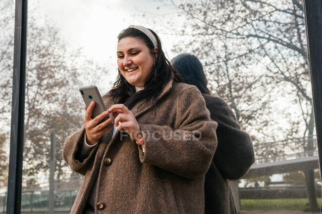 Young glad chubby female in coat surfing internet on mobile phone while reflecting in wall in town — Stock Photo