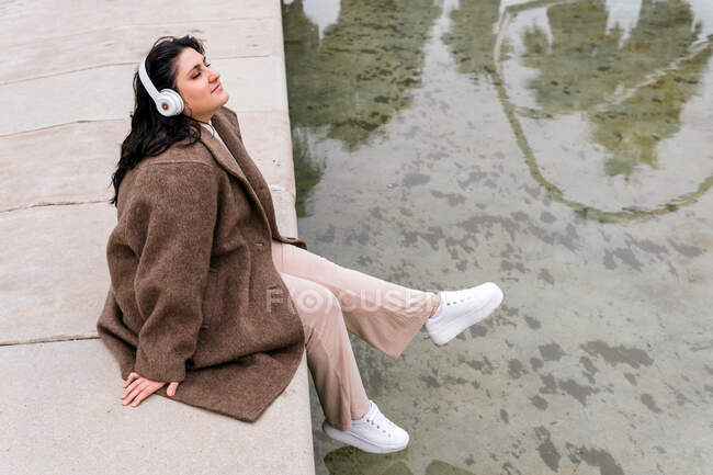 Visão lateral de alto ângulo de sonhador plus size feminino ouvindo música de fones de ouvido sem fio enquanto descansa com os olhos fechados acima do canal urbano — Fotografia de Stock