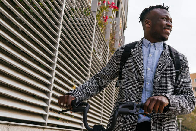 Jovem conteúdo afro-americano masculino empregado no casaco com bicicleta de pé no pavimento urbano contra a parede com nervuras e olhando para longe — Fotografia de Stock