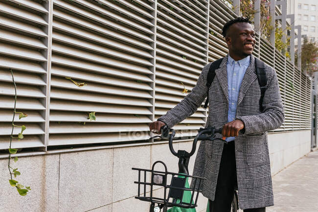Junge zufriedene afroamerikanische männliche Angestellte im Mantel mit Fahrrad steht auf städtischem Bürgersteig gegen gerippte Wand und schaut weg — Stockfoto