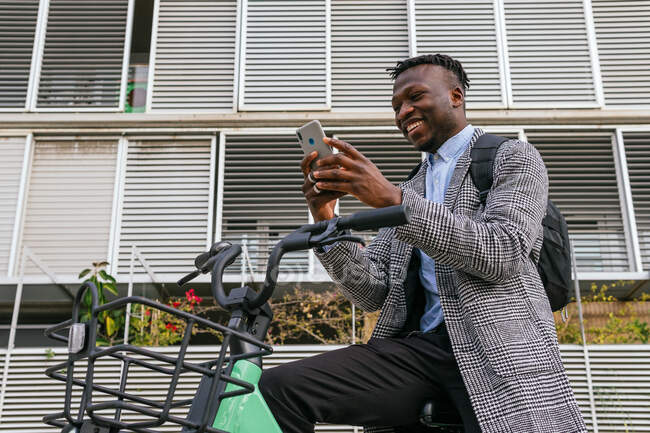 Low angle of content African American male manager writing text messaging on cellphone near bicycle in town — Stock Photo