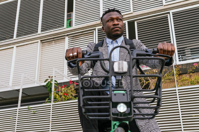 Jovem conteúdo afro-americano masculino empregado no casaco com bicicleta de pé no pavimento urbano contra a parede com nervuras — Fotografia de Stock