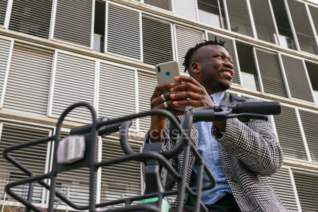 Low angle of content African American male manager writing text messaging on cellphone near bicycle in town — Stock Photo